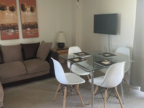 Een moderne woonkamer met een eettafel met glazen blad, witte stoelen, een bruine bank, een lamp en een flatscreen televisie aan de muur.