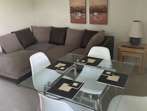 Uma sala de estar moderna com uma mesa de centro de vidro, um sofá em forma de L marrom e uma pequena mesa lateral de madeira com uma luminária.