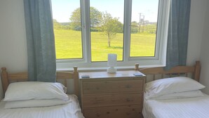Une chambre avec deux lits simples, une commode en bois, une lampe de chevet et une fenêtre donnant sur un champ verdoyant.