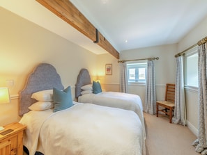 Une chambre avec deux lits, des têtes de lit en bois, une fenêtre avec des rideaux à motifs, une chaise en bois et une table de chevet avec une lampe.