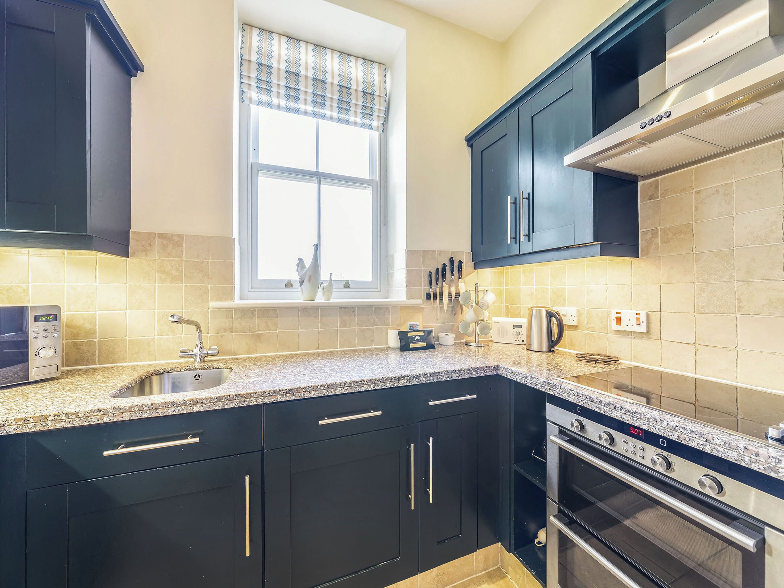 A kitchen with dark cabinets, a window with patterned valance, a microwave, a kettle, and a stove.