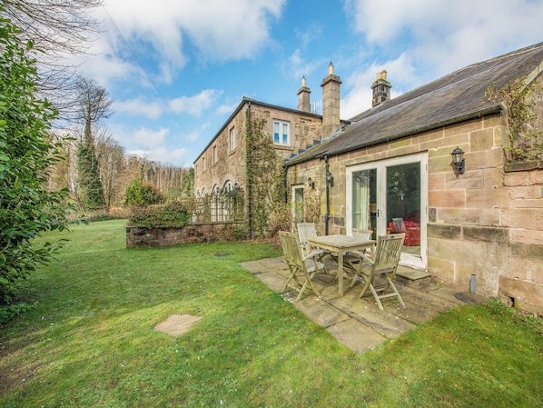 A stone building with a patio, a wooden table and chairs, and a view of the surrounding garden.