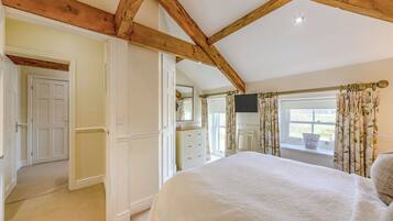 A bedroom with a large bed, wooden ceiling beams, a television, and a window with curtains.