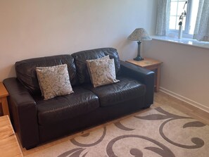 A dark leather sofa with floral cushions, a small wooden side table, a lamp, and a patterned rug.