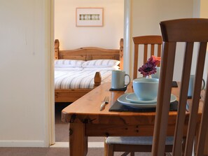 A wooden dining table with a cup, saucer, and a flower arrangement. A bed with striped bedding is visible in the background.