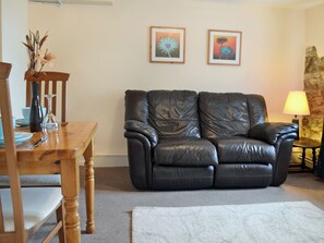 A living room with a leather sofa, wooden dining table, chairs, a vase with flowers, framed pictures on the wall, and a lamp on a side table.