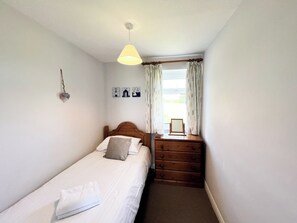 Une petite chambre à coucher, bien rangée, avec un lit simple, une commode en bois, une table de chevet avec une lampe et une fenêtre avec des rideaux.