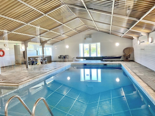 An indoor swimming pool with a tiled floor, a wooden ceiling, and a seating area with chairs.