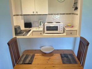 Een kleine keuken met een magnetron, broodrooster en waterkoker, een klein spoelgedeelte en een eettafel met twee stoelen.