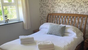 A neatly made bed with white linens and two folded towels, a wooden headboard, and a window with curtains. - Hawfinch Apartment - UK49304 (Woolfardisworthy, Clovelly)
