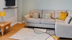 Ein Wohnzimmer mit grauem Sofa, gemusterten Kissen, einem Holztisch, einer Stehlampe und einem Flachbildfernseher an einer Wandhalterung.