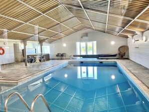 Une piscine intérieure avec un sol carrelé, un plafond en bois et un espace détente avec des chaises.