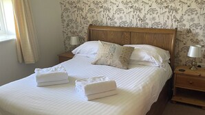 A neatly made bed with white linens and two folded towels, wooden headboard, floral wallpaper, bedside lamps, and a window with curtains. - Chaffinch Apartment - UK49307 (Woolfardisworthy, Clovelly)