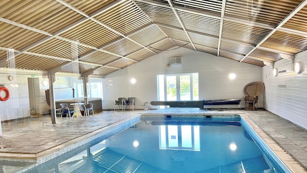 Una piscina interior con suelo de baldosas, techo de madera y un área de descanso con sillas.