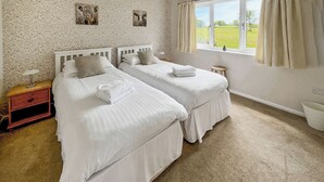 A bedroom with two beds, a nightstand, a window with curtains, and a wall with a floral pattern. - Jasmine Apartment - UK49309 (Woolfardisworthy, Clovelly)