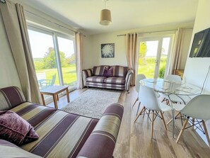 A living room with a sofa, a dining table with chairs, a glass coffee table, and a view of the outdoors. - Jasmine Apartment - UK49309 (Woolfardisworthy, Clovelly)