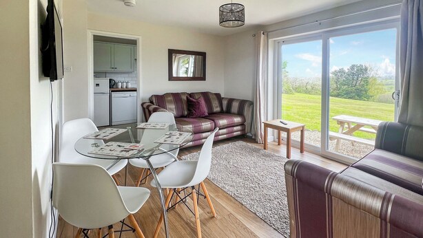 A dining area with a glass table and chairs, a sofa, and a view of the outdoors. - Jasmine Apartment - UK49309 (Woolfardisworthy, Clovelly)