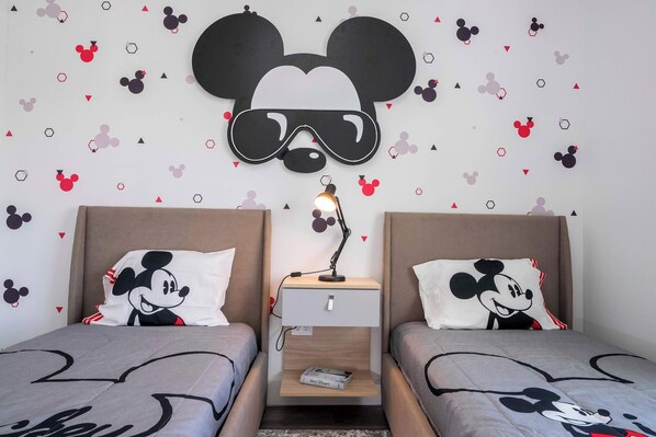 Una habitación con dos camas, cada una con ropa de cama temática de Mickey Mouse, una mesita de noche con una lámpara y una decoración de pared que muestra a Mickey Mouse con gafas de protección.