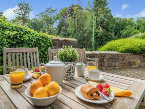 Une table en bois, dressée à l’extérieur, sur laquelle on a disposé des aliments pour le petit-déjeuner, une théière et une tasse de café, avec de la verdure en arrière-plan.