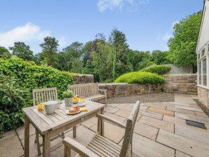 Une table en bois, entourée de chaises et d’un banc, le tout bordé d’un pavage en pierre et de verdure.