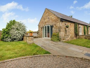 A stone building with a sloped roof, a paved entrance, and a small garden area with a bench and a large bush.