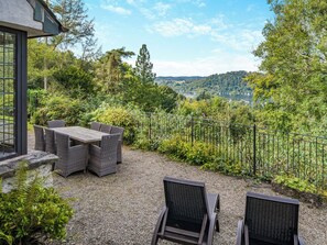 An outdoor patio with a table and chairs, surrounded by gravel and greenery, overlooking a lake and hills. - 4 bedroom accommodation in Windermere (Windermere)