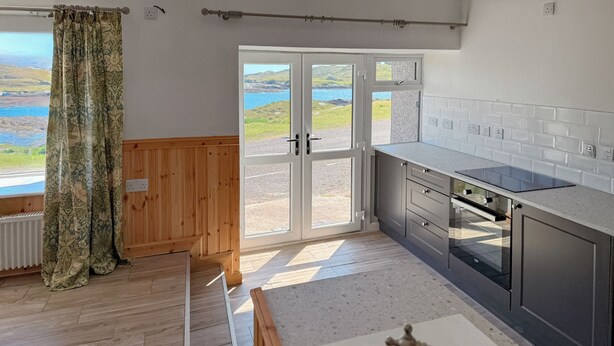 Una cucina moderna con pavimento in legno, piano cottura integrato e vista su un lago attraverso una porta in vetro.