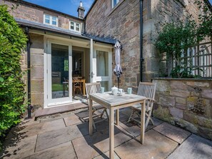 Edificio de piedra con un patio que incluye una mesa y sillas de madera, una ventana y una puerta que da a un espacio interior.