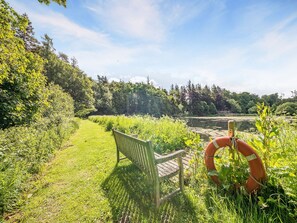 A bench in a grassy area with a lifebuoy nearby, surrounded by greenery and trees.