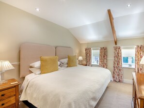 A bedroom with a large bed, a wooden nightstand, a lamp, a window with floral curtains, and a sloped ceiling. - 1 bedroom accommodation in Doxford, near Embleton (Doxford, near Embleton)