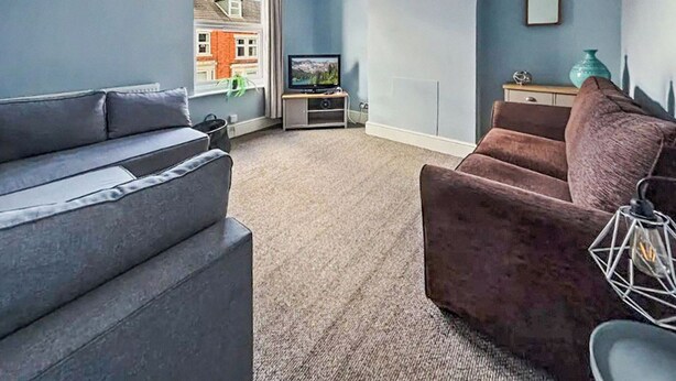 Ein Wohnzimmer mit grauem Sofa, braunem Sessel, Fernseher und Fenster mit Blick auf ein Haus.