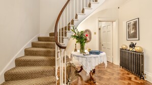 A staircase with patterned carpet, a white table with a vase and a book, and a radiator with decorative spheres.