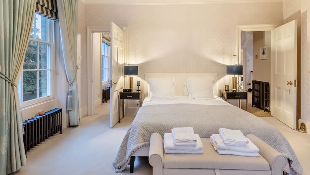 A spacious bedroom with a large bed, bedside tables, a chandelier, and a view through a window with curtains.
