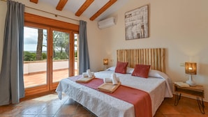 A bedroom with a large bed, wooden beams, a ceiling light, an air conditioning unit, a view through the window, and a painting on the wall.