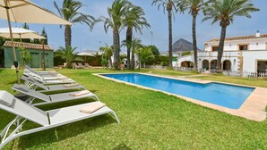 A poolside area with lounge chairs and umbrellas, surrounded by palm trees and a building with arches.