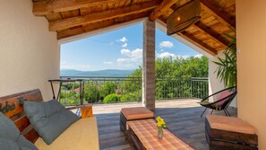 Un balcón con techo de madera, un sofá con cojines, una mesa con un florero y vistas a zonas verdes y montañas.