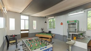 Una habitación con mesa de fútbol, mesa de billar, ventana con persianas, ventilador de techo y cafetera de pared.