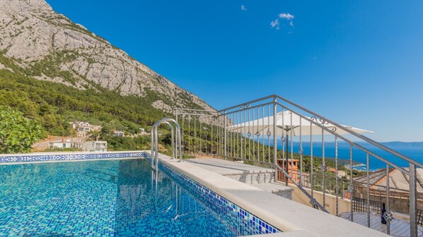 A swimming pool with a tiled edge, a metal staircase leading to a covered area, and a mountainous backdrop.