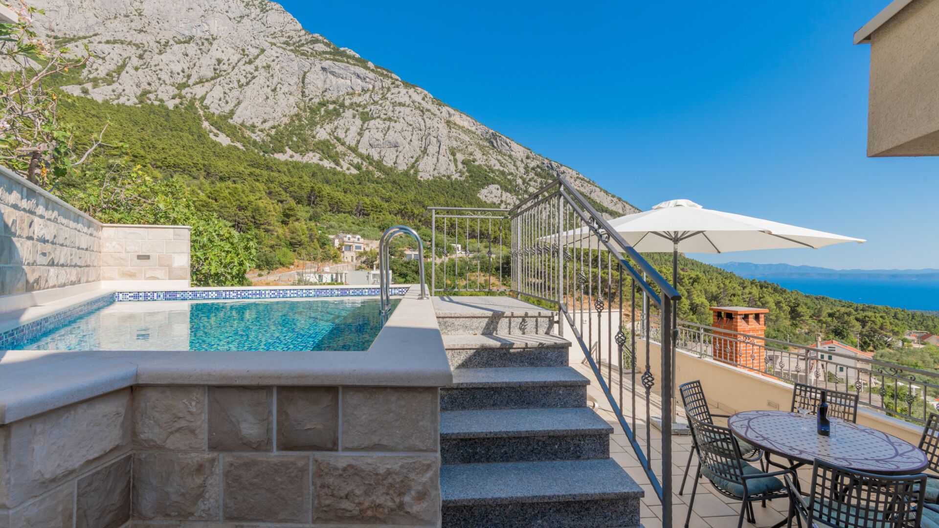 A pool with clear blue water, stone steps leading up to it, a white umbrella, and a view of a mountainous landscape.