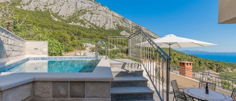 A pool with clear blue water, stone steps leading up to it, a white umbrella, and a view of a mountainous landscape.