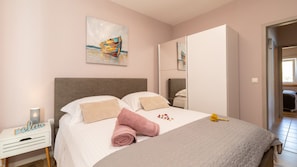 Een slaapkamer met een bed, kussens, een opgerolde handdoek, een nachtkastje, een spiegel en een schilderij aan de muur.