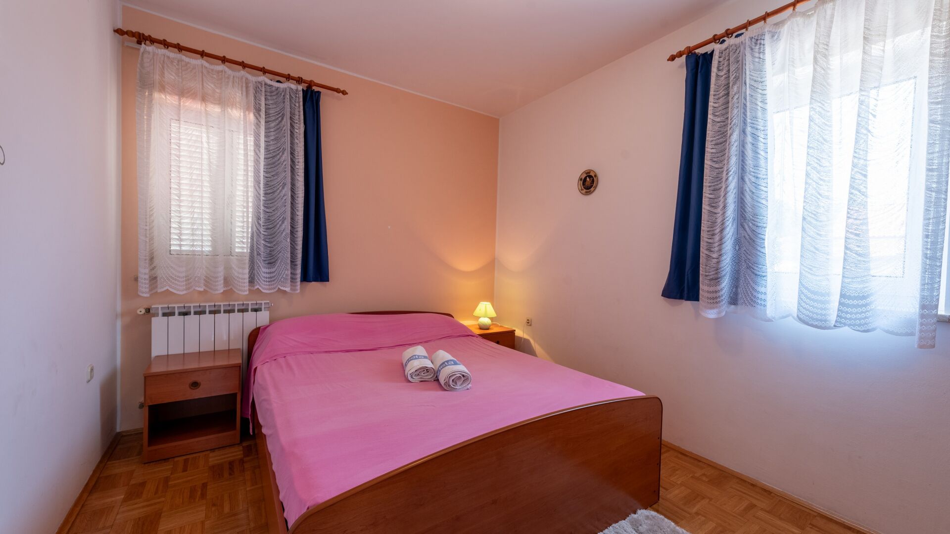 Een slaapkamer met een houten bed, een radiator, een nachtkastje met een lamp en twee ramen met gordijnen.