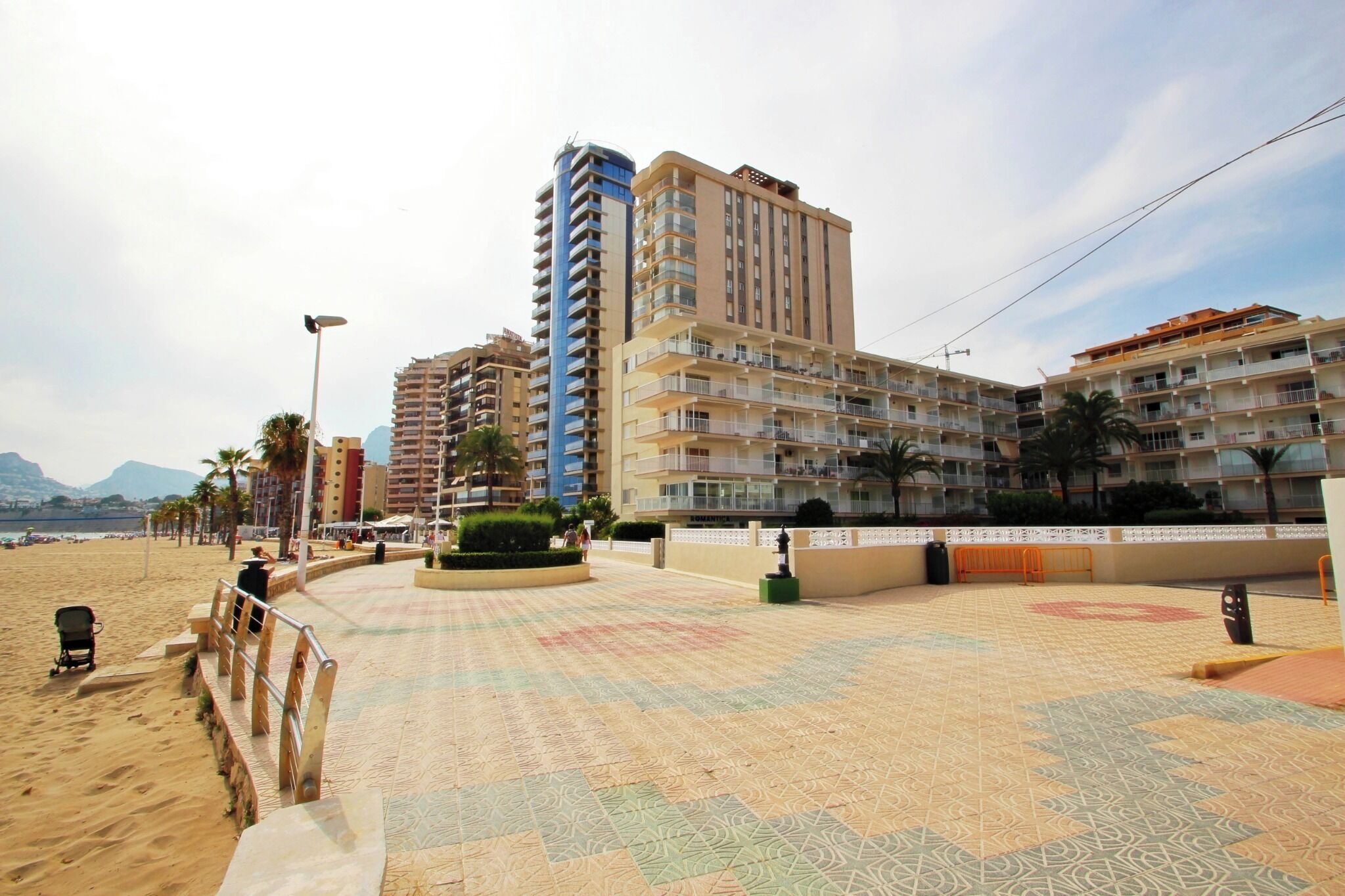 Ein Strandhotel mit mehreren Stockwerken, Balkonen und einem Sandstrand im Vordergrund.