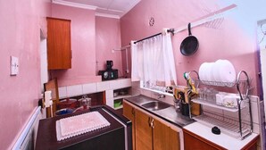 A compact kitchen with pink walls, a wooden cabinet, a microwave, a sink, and a window with a curtain. - Lux Suites The Pink House Naivasha (Naivasha)