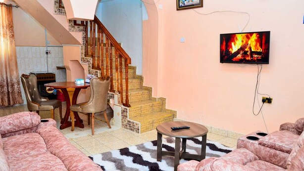 A living room with a staircase, a dining area, and a TV displaying a fire. - Lux Suites The Pink House Naivasha (Naivasha)