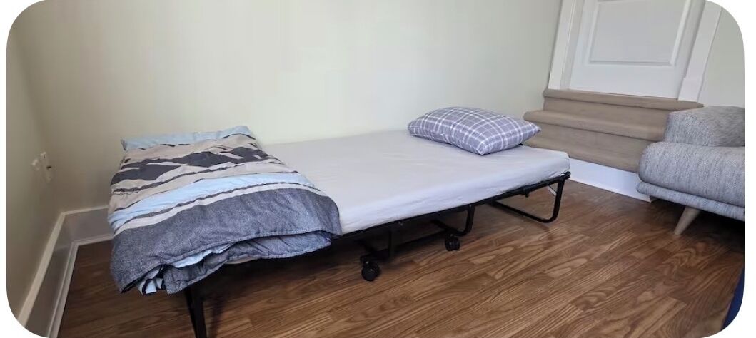Ein kleines Zimmer mit einem Einzelbett, Holzboden, einer grauen Couch und einer Treppe, die in einen oberen Bereich führt.