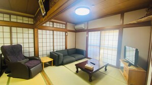 A traditional Japanese-style room with tatami flooring, a tatami mat table, a sofa, a chair, and a television. - Tsukiyama Inn (Yamatotakada)