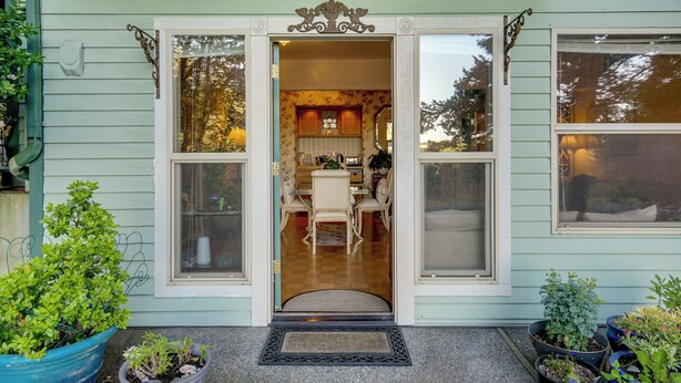 Uma casa com fachada verde, porta frontal com painéis de vidro, área de jantar interna e diversos vasos de plantas do lado de fora.