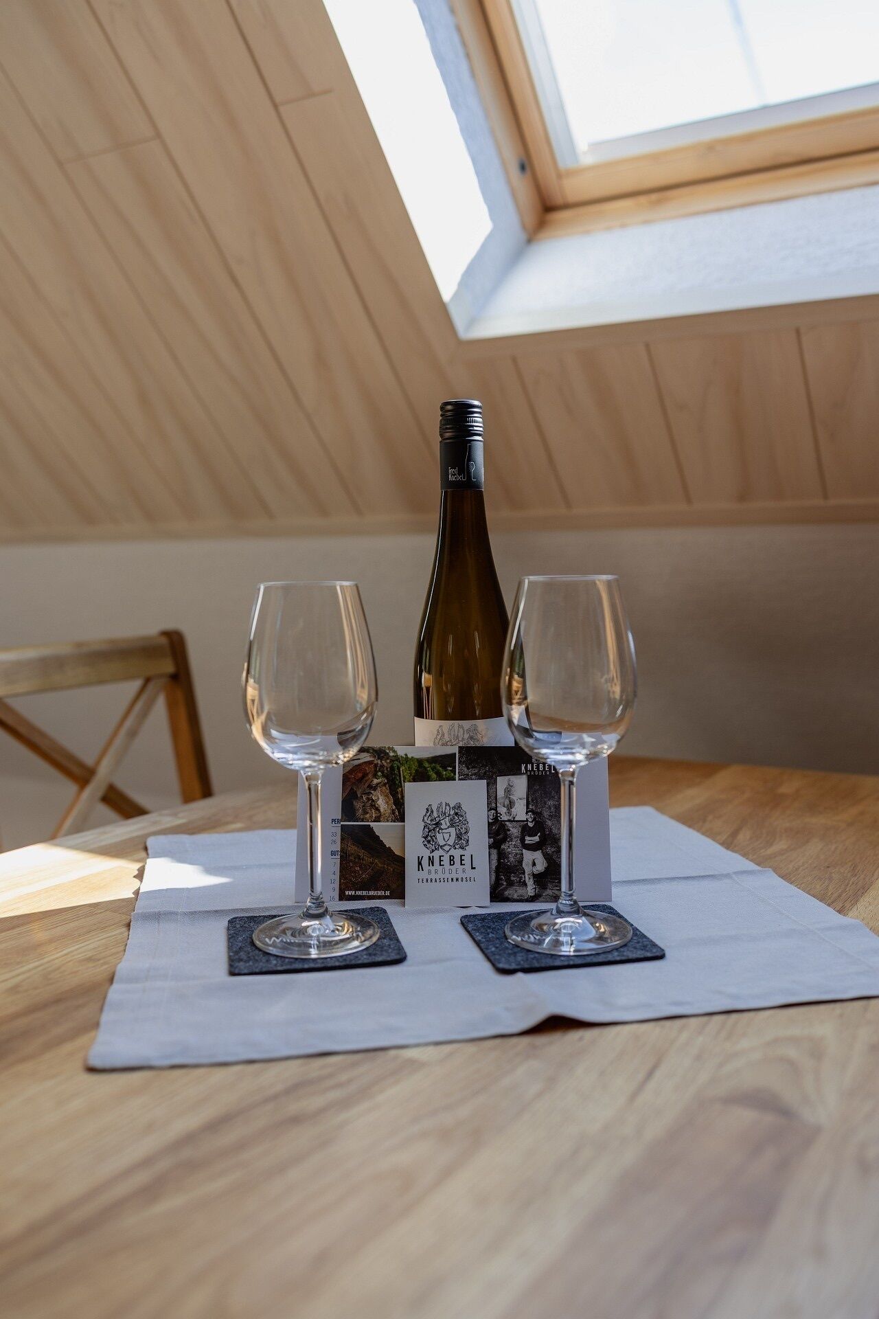 A bottle of wine on a table with two glasses and a placemat, under a skylight.