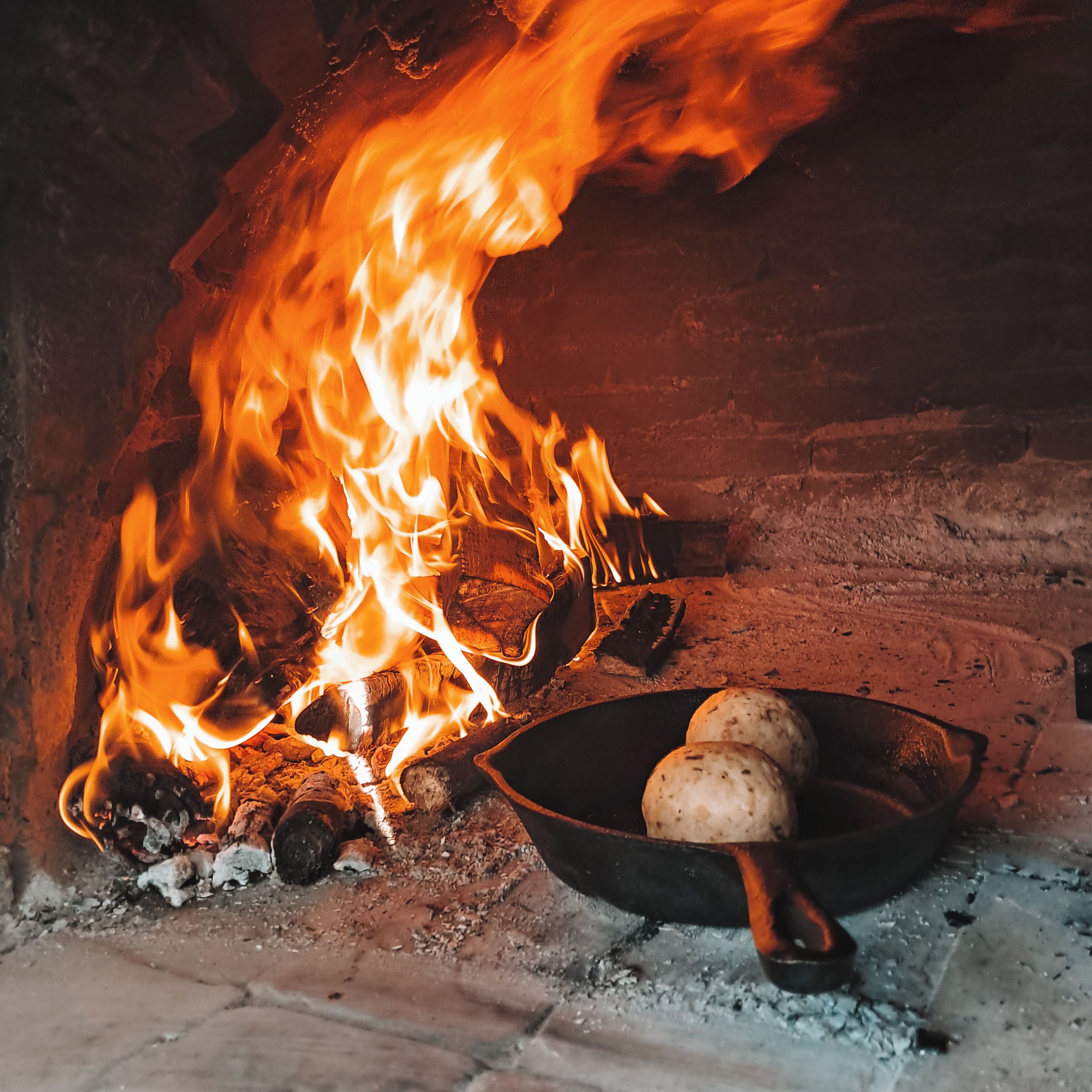 A traditional fireplace with logs and burning flames, a cast iron skillet with bread, and a rustic brick wall.